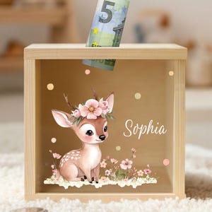 Könnte beinhalten: Eine Holzspardose mit einem Schlitz oben. Die Vorderseite zeigt ein Cartoon-Reh mit einem Blumenkranz und dem Namen "Sophia". Ein Fünf-Euro-Schein steckt teilweise im Schlitz. Die Box hat eine hellbraune Oberfläche.