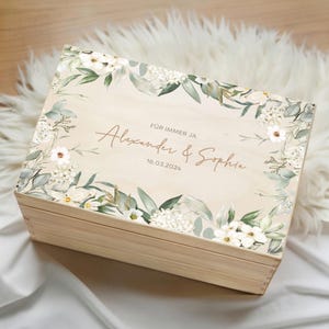 Può includere: Un cofanetto in legno chiaro, decorato con un motivo floreale con fiori bianchi e foglie verdi. Il coperchio presenta la scritta "FÜR IMMER JA" sopra i nomi "Alexander & Sophia" e la data "16.03.2024".