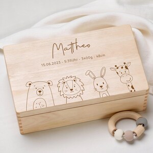 Può includere: Una scatola ricordo in legno chiaro con il nome "Mattheo" e i dettagli della nascita. La scatola è decorata con disegni a tratto di un orso, un leone, un coniglio e una giraffa. Un sonaglio in legno con perline grigie, bianche e rosa è accanto.