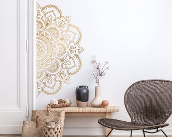 May include: A gold mandala wall decal with intricate details, partially covering a white wall. The decal is placed above a wooden table with a vase of white cotton flowers, a black vase, and a wicker basket.