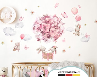 May include: Pink and white watercolor bunny rabbit wall decals with a pink flower hot air balloon, clouds, and butterflies.  The decals are on a white wall with a wooden crib in the foreground.  The text "MADE IN GERMANY" is on the crib.