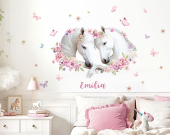 Könnte beinhalten: Zwei weiße Pferde mit rosafarbenen Blumenkränzen und einem Blumenkranz um sie herum. Der Name "Emilia" steht unter den Pferden. Das Bild ist an einer weißen Wand mit einigen rosa Schmetterlingen.
