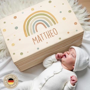 Pode incluir: Uma caixa de madeira clara com um design de arco-íris e o nome "MATTHEO". A caixa é decorada com cores pastel. Um bebê com roupa e chapéu brancos está deitado ao lado.