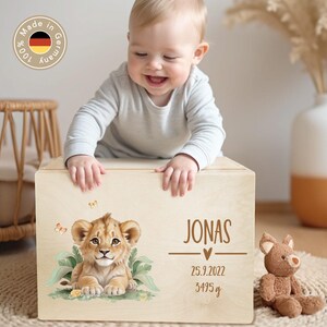 May include: A wooden keepsake box featuring a lion cub illustration, the name "Jonas", birth details, and weight. A baby is smiling and leaning on the box. A stuffed bear sits nearby. The box has a "Made in Germany" sticker.