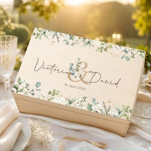 Pode incluir: Uma caixa retangular de madeira clara com um design floral impresso de folhas verdes e flores brancas nas bordas superior e inferior. O centro exibe o texto Victoria & David com a data 16. April 2026 em preto.