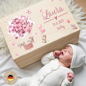 Op de afbeelding: Een lichtgekleurde houten kist met een bloemen- en konijnenontwerp, gepersonaliseerd met de naam "Laura", datum en gewicht. De kist is versierd met roze bloemen, ballonnen en vlinders. Een baby in een witte romper en muts ligt naast de kist.