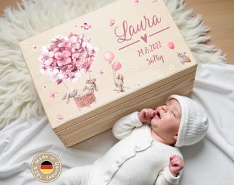 May include: A light-colored wooden box with a floral and bunny design, personalized with the name "Laura", date, and weight. The box is decorated with pink flowers, balloons, and butterflies. A baby in a white onesie and hat is lying next to the box.
