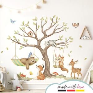 Könnte beinhalten: Ein Wandaufkleber im Aquarellstil mit einem Baum, auf dem ein Bär in einer Hängematte schläft, ein Fuchs, ein Reh und ein Kaninchen und ein Eichhörnchen auf einer Schaukel. Der Aufkleber enthält auch ein Vogelhaus, Schmetterlinge und den Text "Meine Regeln".