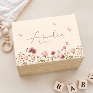 Op de afbeelding: Een rechthoekige, lichtgekleurde houten doos met een bloemenprint van roze en paarse bloemen, groene stengels, een kleine bij en vlinders. De naam Amalia en de datum 11.04.2022 zijn in een donkerroze lettertype op de doos gedrukt.