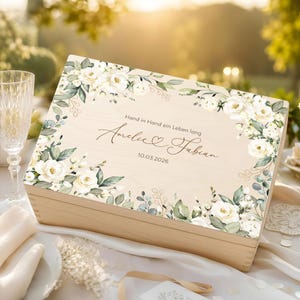 Op de afbeelding: Een rechthoekige houten doos met een bloemenprint van witte rozen en groene bladeren. De tekst luidt: Hand in Hand ein Leben lang, Amelie & Fabian, 10.03.2026. De doos heeft een lichte houtnerf en een scharnierend deksel met een decoratieve rand.