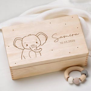 Può includere: Un cofanetto in legno chiaro con un'illustrazione di elefante incisa e il nome "Samira" con la data 10.03.2025. Un sonaglio in legno con perline bianche, rosa e grigie è accanto alla scatola.