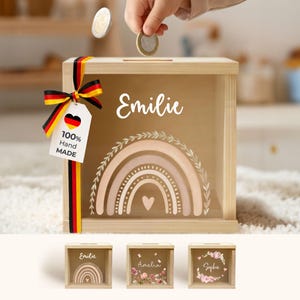 Könnte beinhalten: Eine quadratische Spardose aus Holz mit einer transparenten Vorderseite, die einen weißen Regenbogen und den Namen Emilie zeigt. Ein schwarz-rot-goldenes Band ist seitlich mit einem 100% Hand Made Etikett und einem Herz in Deutschlandfarben befestigt.