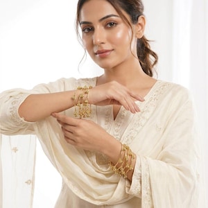 May include: A woman wearing a cream-coloured outfit, with gold bangles on her wrists. The bangles feature a delicate design with small, dangling embellishments. The outfit is made of a soft, flowing fabric.