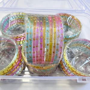 May include: A set of multi-coloured bangles in shades of pink, blue, and yellow, featuring small gold-toned accents. The bangles are arranged in stacks and packaged within a clear plastic storage container with a hinged lid for organisation.