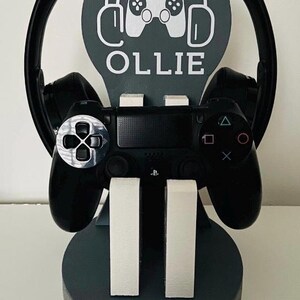May include: A grey wooden stand with a grey and white gamer logo, the name "OLLIE" and a black video game controller and headphones.