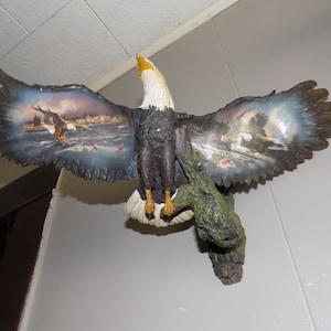Può includere: Una scultura da parete raffigurante un'aquila calva con le ali spiegate, caratterizzata da dettagliati dipinti paesaggistici di aquile in volo sull'acqua. L'aquila ha la testa bianca, il becco giallo, il corpo marrone scuro ed è appollaiata su un ramo d'albero verde e strutturato.