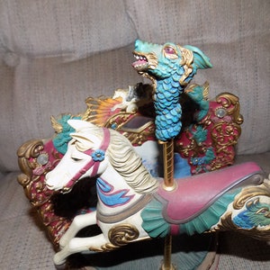 Puede incluir: Un caballo de carrusel blanco con una silla de montar azul y verde y adornos dorados. El caballo forma parte de un carrusel con una cabeza de dragón en un poste.