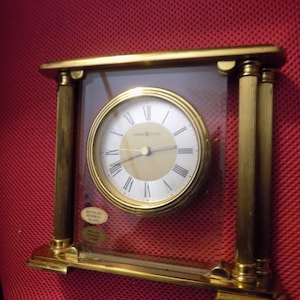 May include: Gold-toned clock with Roman numeral markings on a white face. The clock is encased in a clear glass frame with gold-toned pillars on either side. The clock face reads "Howard Miller". The glass frame has a sticker that reads "Recycled Mineral Glass".