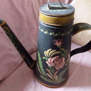 May include: A black metal watering can with a gold trim and painted floral designs in shades of pink, green, and yellow.