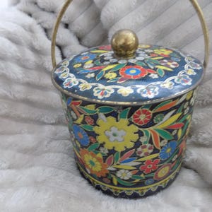 Peut inclure: Une boîte en métal vintage, cylindrique, avec un couvercle et une poignée, ornée d'un motif floral vibrant. Le design présente des fleurs jaunes, rouges, bleues et vertes sur fond bleu foncé. La poignée et le bouton sont dorés.