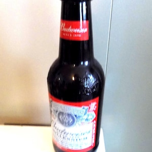 Pode incluir: Uma garrafa de cerveja de vidro castanho escuro com uma tampa prateada. A garrafa apresenta um rótulo de pescoço vermelho com o texto branco Budweiser Since 1876 e um rótulo principal vermelho e branco com o logótipo e texto Budweiser.