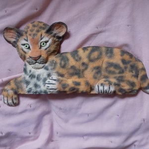 May include: A ceramic figurine of a leopard cub lying down. The cub is brown and black with spots and has green eyes. The figurine is on a pink background.