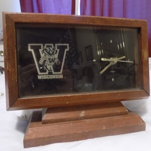 Puede incluir: Un reloj de madera con una esfera negra y un logotipo plateado de la mascota del Wisconsin Badger. La esfera del reloj también tiene una 'W' plateada y la palabra 'WISCONSIN' debajo de la mascota. El reloj está sobre una base de madera.