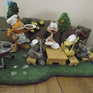 May include: A whimsical figurine depicting a picnic scene with cartoon characters. Garfield, Odie, and other characters are gathered around picnic tables and a grill. The figurine is set on a green base with a wooden fence backdrop.