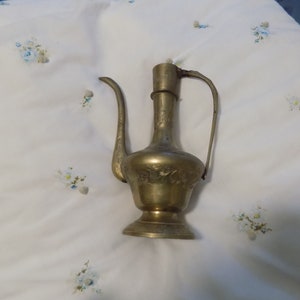 May include: A brass pitcher with an ornate design and a curved handle. The pitcher has a bulbous body and a narrow neck.
