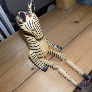 Puede incluir: Figura de cebra de madera con rayas negras y beige. La cebra está sentada con las patas extendidas. La figura tiene orejas negras y una melena negra y marrón. La cebra está sobre una superficie de madera.