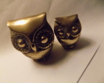 May include: Two brass-colored owl figurines. The owls have detailed facial features, including large, round eyes and textured feathers. The owls are in a decorative style, and are likely used as home decor or collectibles.