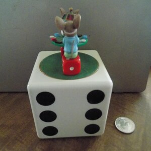 May include: A white ceramic cube shaped like a six-sided die with black pips. On top sits a green circular platform with a small mouse figurine wearing a blue shirt, seated at a table with game pieces, perched on a red die next to a silver coin.
