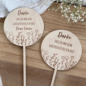 Könnte beinhalten: Zwei runde Pflanzenstecker aus hellem Holz mit dünnen Stielen. Jeder zeigt den lasergravierten deutschen Text "Danke DASS DU MIR BEIM WACHSEN GEHOLFEN HAST" und florale Linienzeichnungen. Ein Stecker enthält zusätzlich den Namen "Deine Emilia".