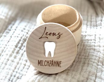May include: A small, round, wooden keepsake box with a lid. The lid features the name "Leong" in cursive above a tooth graphic and the word "MILCHZÄHNE" below. The box is open, revealing its interior.