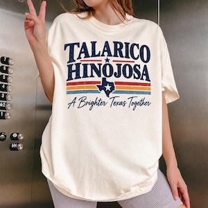 May include: A cream-colored t-shirt featuring the dark blue text Talarico Hinojosa above a graphic of the state of Texas with a star. Below are horizontal red, orange, and blue stripes and the text A Brighter Texas Together in blue.