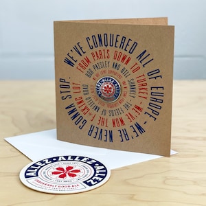 May include: A brown card with a blue and red circular design that says "We've conquered all of Europe from Paris down to Turkey. We've won the f*cking lot. We're never gonna stop."  The card has a small metal pin with a red cross on it. The card is sitting on a white envelope with a round sticker that says "Allez Allez" in blue and red.