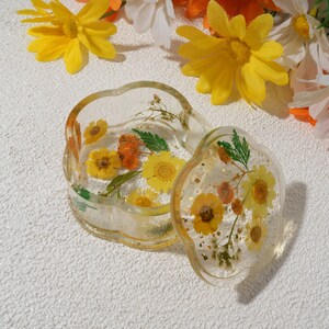 K&ouml;nnte beinhalten: Eine transparente Schmuckschatulle aus Harz mit Blumenmuster. Die Schachtel ist blumenf&ouml;rmig und hat einen Deckel. Deckel und Schachtel sind mit gepressten Blumen in Gelb-, Orange- und Gr&uuml;nt&ouml;nen gef&uuml;llt.