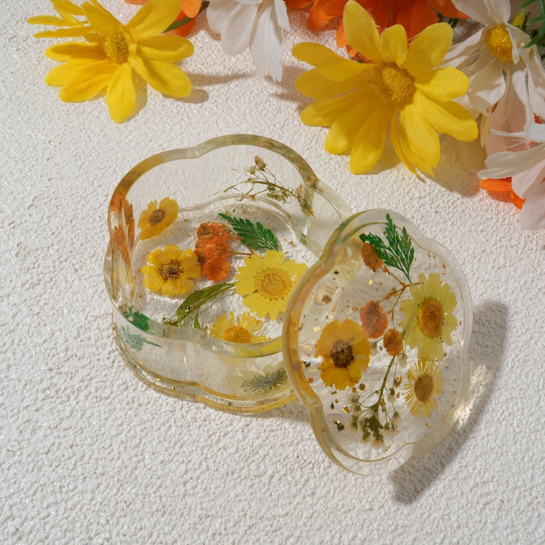 K&ouml;nnte beinhalten: Eine transparente Schmuckschatulle aus Harz mit Blumenmuster. Die Schachtel ist blumenf&ouml;rmig und hat einen Deckel. Deckel und Schachtel sind mit gepressten Blumen in Gelb-, Orange- und Gr&uuml;nt&ouml;nen gef&uuml;llt.