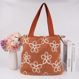 May include: A brown fuzzy tote bag with white embroidered flowers. The bag has brown straps and a drawstring closure.