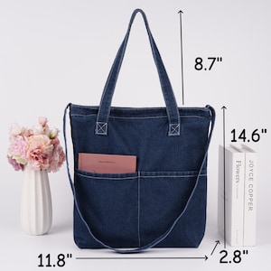Peut inclure: Un sac fourre-tout en jean bleu fonc&eacute; avec une bandouli&egrave;re et deux poches ext&eacute;rieures. Le sac mesure 30 cm de large, 37 cm de haut et 7 cm de profondeur. Les anses mesurent 22 cm de long. Un livre rose est dans la poche.