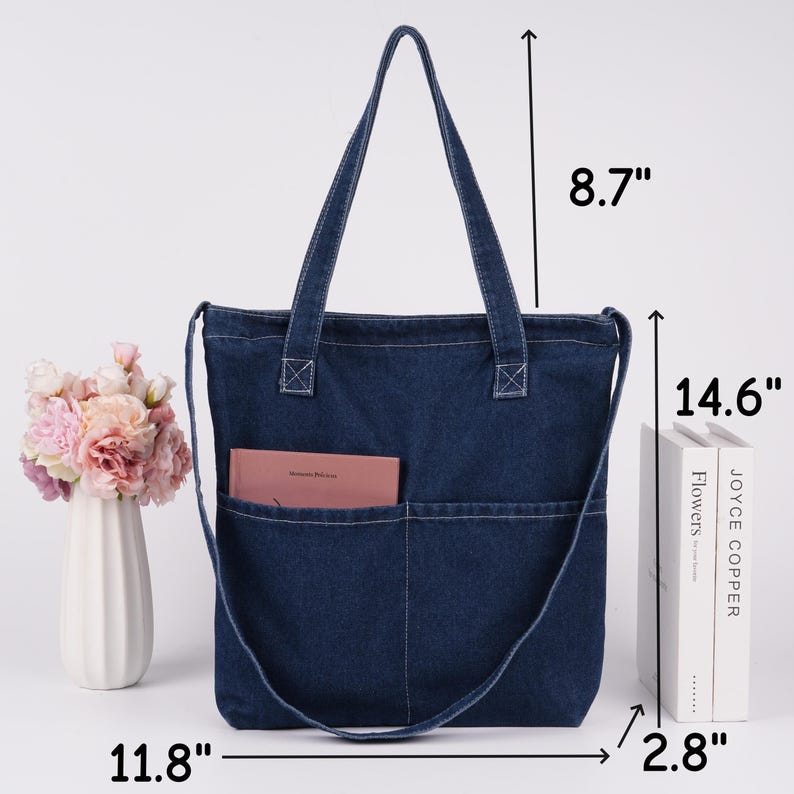 Peut inclure: Un sac fourre-tout en jean bleu fonc&eacute; avec une bandouli&egrave;re et deux poches ext&eacute;rieures. Le sac mesure 30 cm de large, 37 cm de haut et 7 cm de profondeur. Les anses mesurent 22 cm de long. Un livre rose est dans la poche.