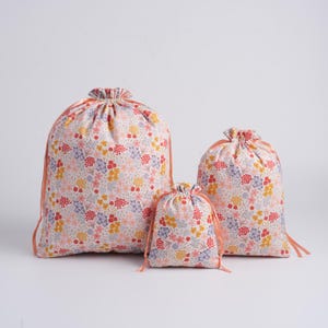 May include: Three drawstring bags of varying sizes made from a light-colored fabric featuring a dense, multi-colored floral pattern in shades of red, yellow, and purple. Each bag is cinched at the top with a matching peach-colored ribbon tie.