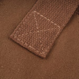 Pode incluir: Uma vista aproximada de uma al&ccedil;a de tecido castanho fixada numa base de lona castanha a condizer. A al&ccedil;a est&aacute; presa com uma costura quadrada com um refor&ccedil;o em forma de X no centro, usando fio de cor clara sobre o material texturizado.