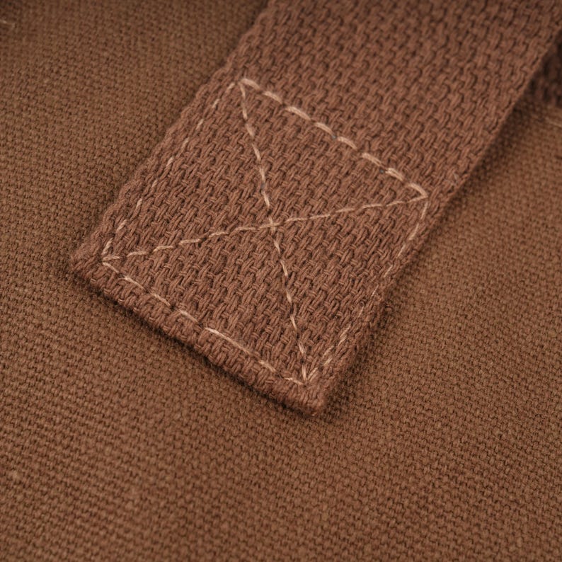 Pode incluir: Uma vista aproximada de uma al&ccedil;a de tecido castanho fixada numa base de lona castanha a condizer. A al&ccedil;a est&aacute; presa com uma costura quadrada com um refor&ccedil;o em forma de X no centro, usando fio de cor clara sobre o material texturizado.