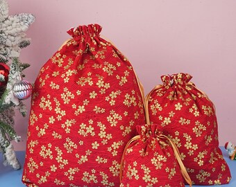 May include: Three red drawstring gift bags with a gold floral pattern. The bags vary in size, with gold ribbon drawstrings. The bags are on a light blue surface, with a Christmas tree and ornaments in the background.