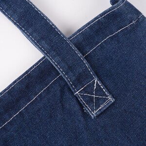 Peut inclure: Gros plan sur un tablier en jean avec des coutures blanches. Les bretelles se croisent dans le dos et sont fix&eacute;es par une couture renforc&eacute;e. Le tablier est de couleur indigo fonc&eacute;.