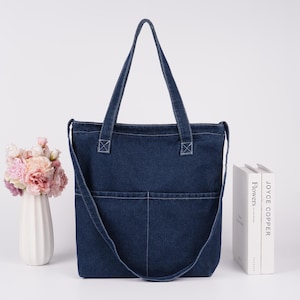 Peut inclure: Un sac fourre-tout en jean bleu fonc&eacute; avec deux poches avant et une longue bandouli&egrave;re. Le sac a deux poign&eacute;es et des coutures blanches. Un vase blanc avec des fleurs roses et p&ecirc;che est &agrave; gauche, et deux livres &agrave; droite.