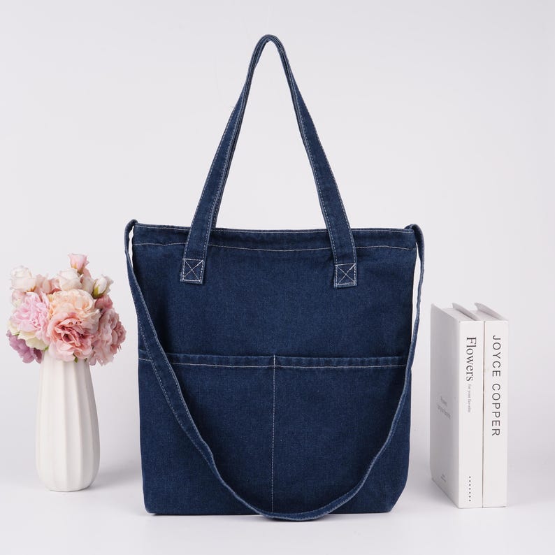 Peut inclure: Un sac fourre-tout en jean bleu fonc&eacute; avec deux poches avant et une longue bandouli&egrave;re. Le sac a deux poign&eacute;es et des coutures blanches. Un vase blanc avec des fleurs roses et p&ecirc;che est &agrave; gauche, et deux livres &agrave; droite.