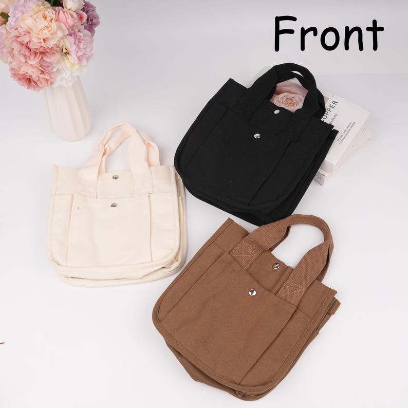 Pode incluir: Tr&ecirc;s pequenas bolsas tote de lona nas cores creme, preta e castanha. Cada bolsa possui duas al&ccedil;as, um bolso frontal com fecho de bot&atilde;o de press&atilde;o em metal prateado e um compartimento principal. A palavra Front est&aacute; impressa a preto no canto.