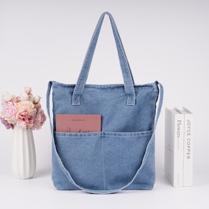 Peut inclure: Un sac fourre-tout en jean bleu clair avec une poche avant et une bandouli&egrave;re. Le sac a deux poign&eacute;es et contient un livre rose. Un vase blanc avec des fleurs roses et blanches est &agrave; gauche, et deux livres &agrave; droite.